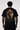 A man viewed from behind wearing a black t-shirt with a large, detailed graphic print on the back. The graphic features a golden, fiery, and dark illustration of a figure with a halo, with Greek and English text below reading 'ΟΤΤΕΝΑΙ ΓΤΩΡΟΜΑΙ ΓΙΝΕΤΑΙ ΣΤΟΥΣ - SPARTANS SPORTWEAR - EST.